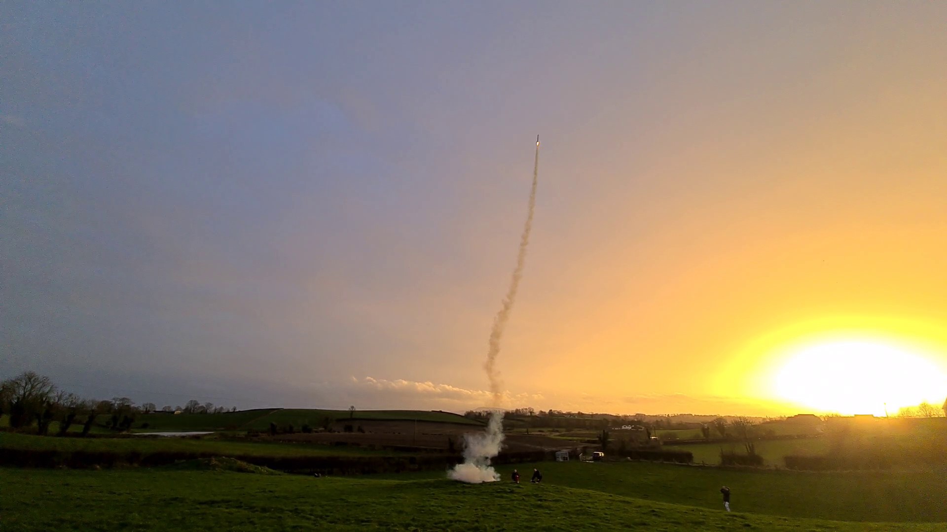 Model rocket launching from field with sunset in background